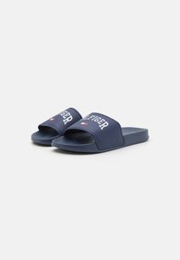 Chanclas de color azul marino con una tira ancha, que presentan la marca "TOMMY HILFIGER" en blanco y un pequeño logo rojo y blanco en el costado. Plantilla texturizada.