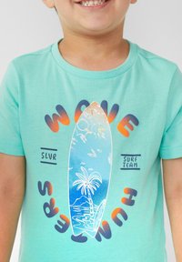 Turquoise katoenen T-shirt met een surfplankgrafiek, een strandscène, een palmboom en de tekst "WAVE HUNTERS" in een verlooplettertype.