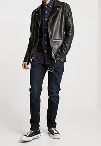 Giacca da motociclista in pelle nera con accessori in argento, indossata sopra una camicia a quadri scura, abbinata a jeans scuri e sneakers nere.
