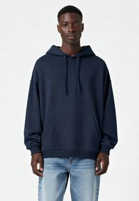 Námornícky modrá mikina s kapucňou vo voľnom strihu, s rebrovanými manžetami a prednou klokankou. Tkanina vyzerá hladko a má textúru.