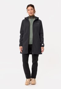 Lange zwarte softshell jas met een capuchon, met een ritssluiting aan de voorkant, gecombineerd met een lichtgroene top en zwarte broek. Lichtgewicht, duurzaam materiaal.