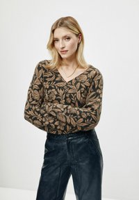 Brązowy sweter w kwiatowy wzór z dekoltem w kształcie litery V, zestawiony z ciemnymi skórzanymi spodniami. Modelka ma proste włosy i prostą złotą naszyjnik.