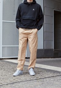 Felpa nera con cappuccio e tasca frontale, abbinata a pantaloni beige e sneakers grigie, in piedi su un marciapiede di ciottoli.