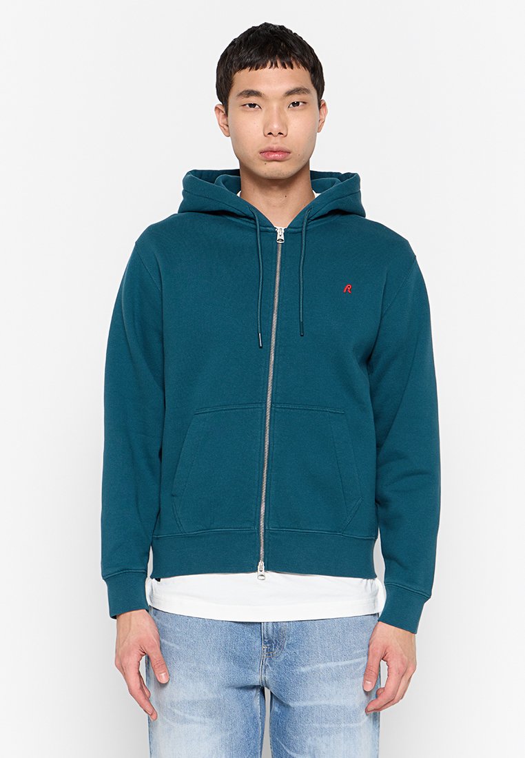 Junger Mann in einem türkisfarbenen Zip-Hoodie mit Kängurutaschen, weißem Unterhemd und hellblauen Jeans, der vor einem weißen Hintergrund steht.