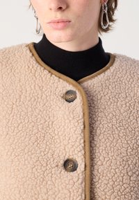 Μπέζ φλις μπουφάν με υφασμάτινη επιφάνεια, καφέ λεπτομέρειες και κουμπί κλεισίματος. Φορεμένο πάνω από μια μαύρη τσέπη, ενισχύει την εναλλασσόμενη εμφάνιση.