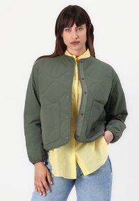 Quilted grøn jakke med knaplukning, rund krave og to forlommer, båret over en lys gul skjorte med knapper, parret med blå jeans.