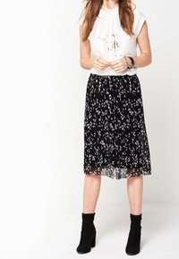 Bílý top s krátkým rukávem a jemným potiskem, zkombinovaný s černou plisovanou sukní s bílým abstraktním vzorem, a černými kotníkovými botami.