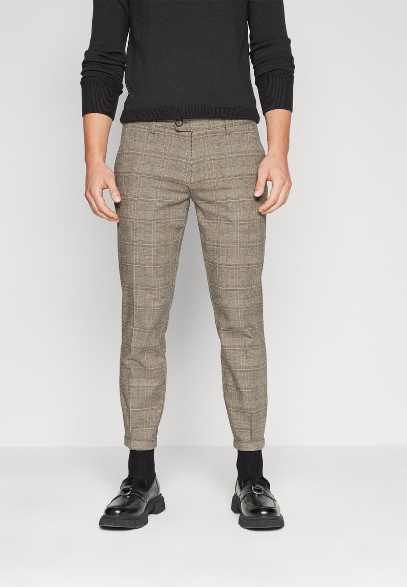 Pantalons ajustés à motifs en plaid marron et beige, avec une taille plate, des poches latérales et une fermeture à bouton, associés à des chaussures et des chaussettes noires.
