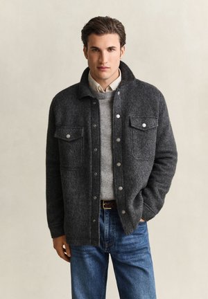 Jeune homme portant une veste en laine gris foncé boutonnée par-dessus un pull gris et une chemise blanche, avec un jean bleu et une main dans la poche.