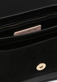 Borsa in pelle nera con chiusura magnetica in metallo dorato e etichetta interna rosa con la scritta "Coccinelle" in rilievo. Tasca con cerniera visibile.