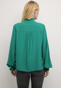 Blusa verde con colletto arricciato, maniche lunghe con polsini elastici, yoke posteriore arricciato e tessuto leggero e morbido.