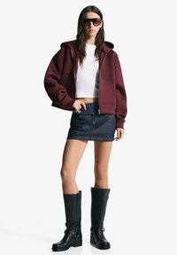 Bordeaux zip-up hoodie, witte cropped top, blauwe denim minirok, kniehoge zwarte laarzen met gespen, en grote zwarte zonnebril.