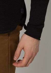 Svart stickad tröja med rundad nederkant, med en sidetikett. Hand delvis synlig, vilande nära ljusbruna byxor. Slät textur.