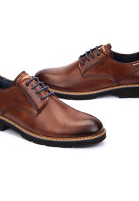 Scarpe in pelle marrone con una texture liscia, dotate di lacci blu e cuciture beige a contrasto lungo la suola. Design classico, punta arrotondata.