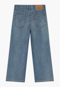 Vaalean siniset leveälahkeiset farkut, joissa on haalistunut viimeistely, ruskea nahkaleima ja kaksi taka-taskua, joissa on allekirjoitusompelu.