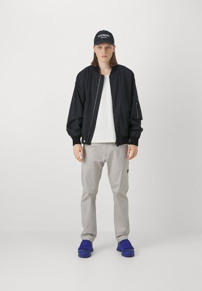 Giubbotto bomber nero con polsini a costine, t-shirt bianca, pantaloni cargo grigio chiaro e sneakers blu. Cappellino nero con testo "ICEBERG".
