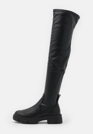 Botte noire au-dessus du genou avec semelle épaisse, bout arrondi et languette de traction à l'arrière, présentée sur un fond blanc uni.