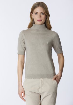 Donna con capelli castano chiaro che indossa un maglione a collo alto beige a maniche corte e pantaloni chiari, con una mano in tasca, su uno sfondo bianco.