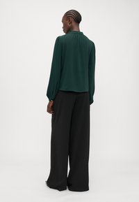 Donkergroen blouse met lange mouwen, een gerimpelde kraag en plooien aan de achterkant, gedragen met een brede zwarte broek. Super gladde stof.
