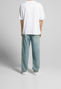 Persona vestida con una camiseta blanca sencilla, jeans holgados de color azul claro y zapatillas blancas, de pie de espaldas contra un fondo neutro.