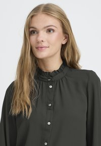 Dunkelgrüne Bluse mit Rüschensaum, Knopfverschluss vorne und langen Ärmeln, aus einem glatten Stoff mit leichtem Glanz.