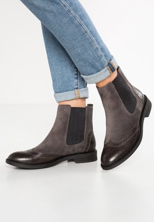 Botas Chelsea de ante gris con detalles de brogue en cuero oscuro, usadas con vaqueros azules remangados sobre un fondo liso.