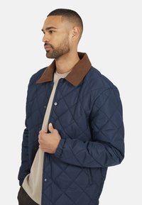 Marineblauwe gewatteerde jas met een bruine corduroy kraag, voorzien van drukknoopsluitingen, lange mouwen en een lichtgewicht ontwerp. Textuur oppervlakten en naden.