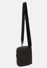 Schwarze Nylon-Crossbody-Tasche mit strukturierten Oberflächen, Reißverschluss, verstellbarem Schultergurt mit Logo und einem runden Emblem auf der Vorderseite.