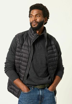 Mann mit Bart, der eine schwarze Steppweste über einem grauen Pullover und blauen Jeans trägt, steht mit Händen in den Taschen und schaut leicht zur Seite.