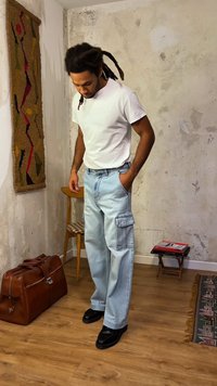 Άνδρας με ντέρεκλους που φοράει λευκό μπλουζάκι, ανοιχτό μπλε cargo τζιν, μαύρα παπούτσια, στέκεται μέσα σε εσωτερικό χώρο κοντά σε ξύλινη καρέκλα και καφέ δερμάτινη τσάντα.