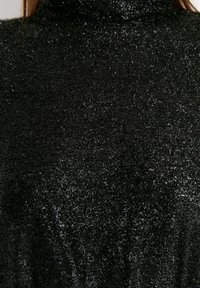 Haut à col roulé noir en tissu texturé et scintillant. La surface est couverte de fils chatoyants, créant une apparence brillante.