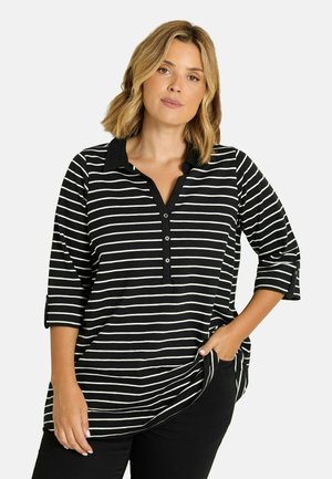 Mujer con cabello rubio hasta los hombros que lleva una camisa de manga tres cuartos a rayas negras y blancas con botones y pantalones negros.