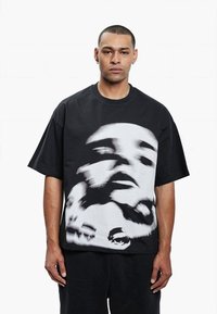 Černé oversize tričko vyrobené z bavlny, které má uprostřed velký abstraktní monochromatický grafický motiv tváře. Krátké rukávy a kulatý výstřih.