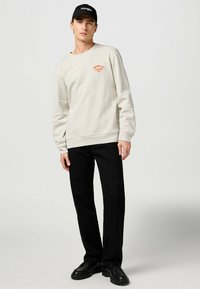 Jasnoszary sweter z pomarańczowym logo "Wrangler Jeans", okrągły dekolt, ściągacze przy rękawach, w zestawieniu z czarnymi spodniami i czarnymi butami.