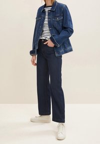 Giacca di jeans, blu scuro, con due tasche frontali e chiusura a bottoni. Camicia a righe sotto. Jeans larghi scuri e sneakers bianche.