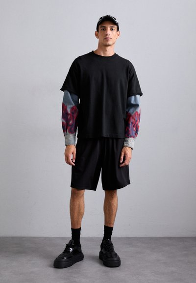 Tricou negru cu mâneci cu model de flăcări gri și roșii, asociat cu pantaloni scurți negri. Adidași negri și o șapcă neagră completează ținuta.
