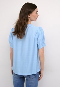 Jasnoniebieska bluzka z krótkim rękawem, z marszczonymi ramionami, okrągłym dekoltem i luźnym krojem, połączona z dżinsami. Gładki materiał, zwiewny fason.