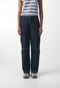 Pantalones cargo de mezclilla oscura, con bolsillos frontales y laterales, corte holgado y costuras visibles. Combinados con una camisa de rayas de manga corta.