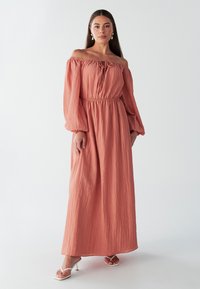 Didelės apimties koralinės spalvos maxi suknelė su tekstūruota medžiaga, baliono formos rankovėmis, elastinga juosmens dalimi ir priekinio raištelio detale.