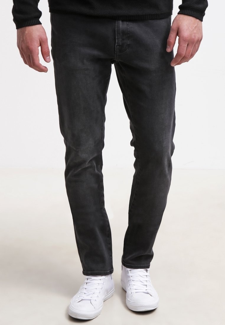 Jean slim gris foncé avec une texture lisse, doté de poches avant et de passants pour ceinture, assorti à des baskets blanches.