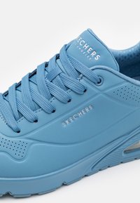 Sapatilha azul clara com detalhes em malha, sola de borracha texturizada, detalhes perfurados e logo "SKECHERS" em branco na lingueta.