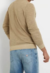 Uomo che indossa un maglione lavorato a maglia beige e jeans azzurri, con un piccolo logo triangolare sulla tasca posteriore, rivolto di spalle rispetto alla fotocamera.