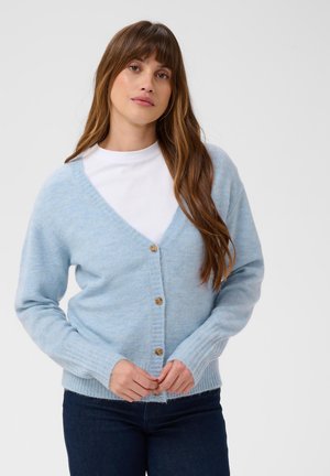 Femme aux longs cheveux bruns porte un cardigan bleu clair boutonné sur un haut blanc et un jean foncé, debout devant un fond uni.