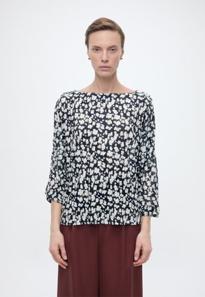 Frau mit kurzen Haaren, die goldene Ohrringe und eine schwarz-weiße Bluse mit Blumenmuster und Dreiviertelärmeln sowie bordeauxrote Hosen trägt, steht vor einem weißen Hintergrund.