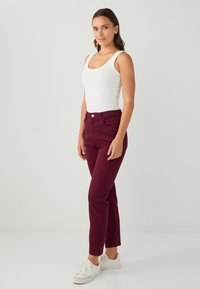 Jeans bordeaux con un design aderente, dotati di chiusura frontale con bottone e tasche, abbinati a una canotta bianca e sneakers bianche.