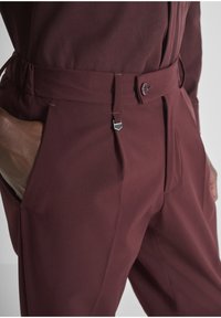 Bordeaux kleurige op maat gemaakte broeken hebben een gladde textuur, zijzakken, een knoopsluiting en een metalen clipaccent op de tailleband.