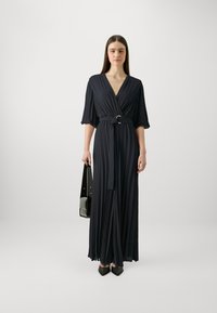 Abito maxi a pieghe di colore navy con scollatura a V profonda, vita cinchia e maniche larghe. Abbinato a una borsa nera e grigia e scarpe nere appuntite.