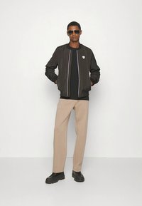 Man som bär svart bomberjacka, svart tröja, beige byxor, svarta skor och mörka solglasögon, stående med händerna i fickorna.