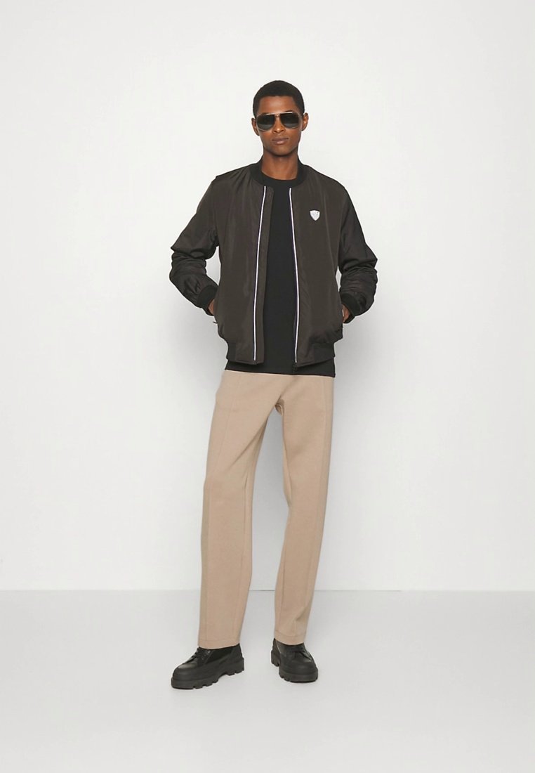 Homme portant une veste bomber noire, une chemise noire, un pantalon beige, des chaussures noires et des lunettes de soleil foncées, debout avec les mains dans les poches.