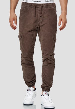 Pantalones cargo marrones con puños elásticos y cinturilla con cordón; cuenta con bolsillos laterales y una tela suave y texturizada. Combinados con zapatillas blancas.